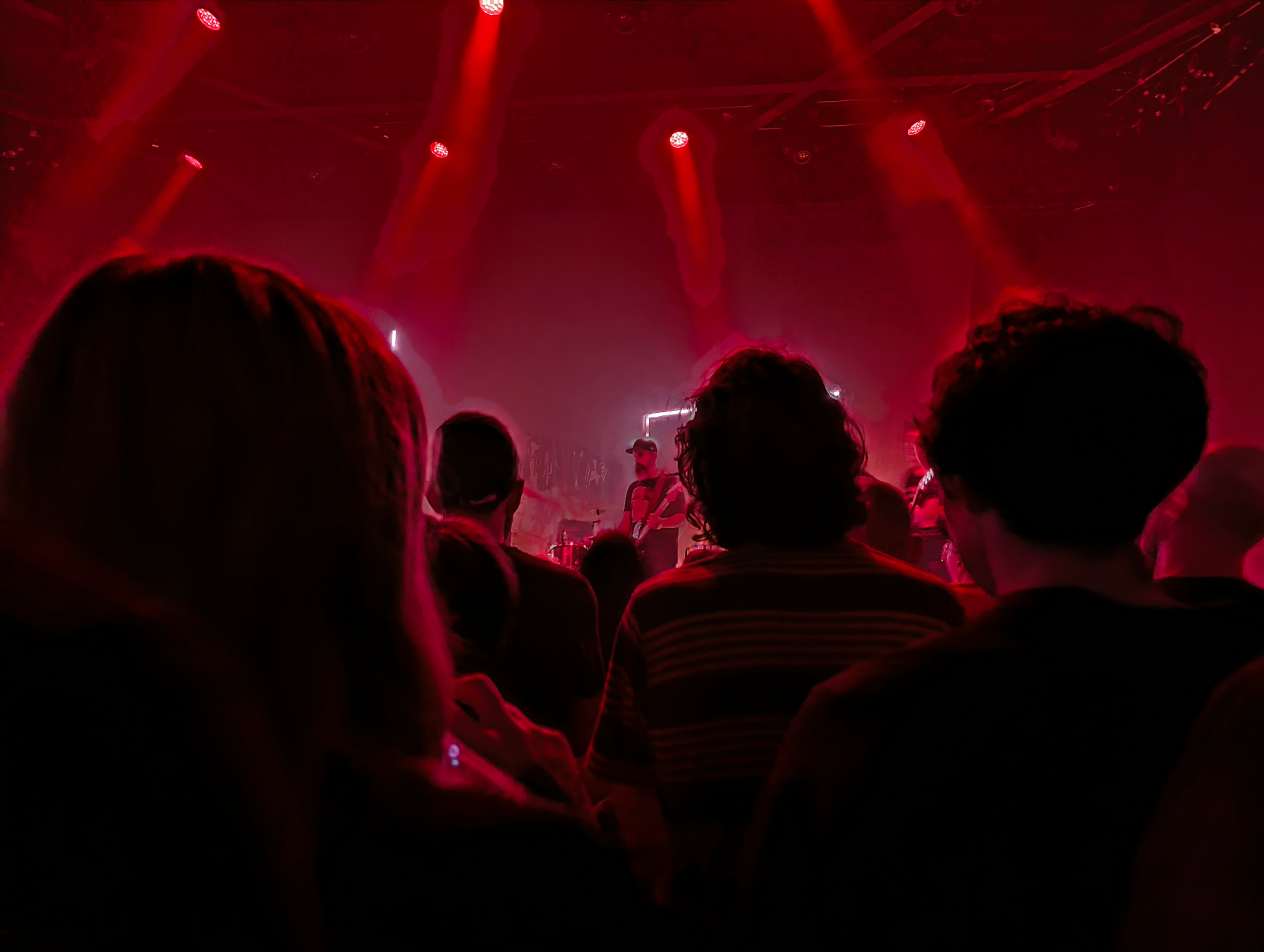 A crowd seen from behind at a live gig, bathed in deep red stage light, watching a band perform